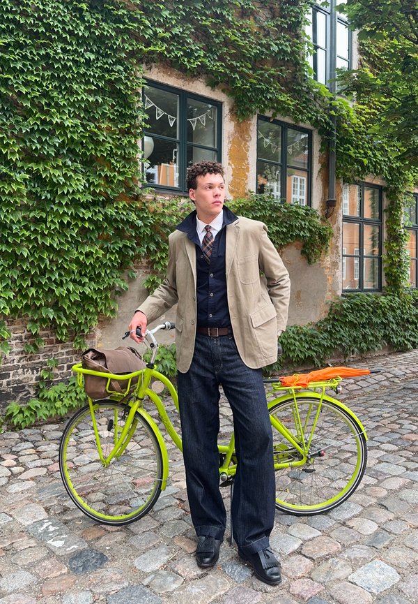 Jonge man in een beige jas en stropdas staat naast een groene fiets op een geplaveide straat met met klimop bedekte muren en ramen.