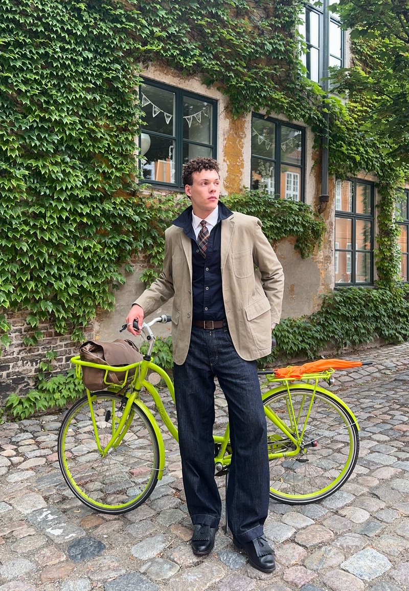 Νέος άνδρας με μπεζ σακάκι και γραβάτα, στέκεται δίπλα σε ένα πράσινο ποδήλατο σε έναν πλακόστρωτο δρόμο με τοίχους και παράθυρα καλυμμένα με κληματαριά.
