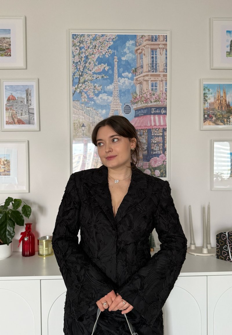 Blazer noir à texture florale avec manches larges, doté d'un décolleté en V profond. Accessoires comprenant des colliers superposés et un sac à main.