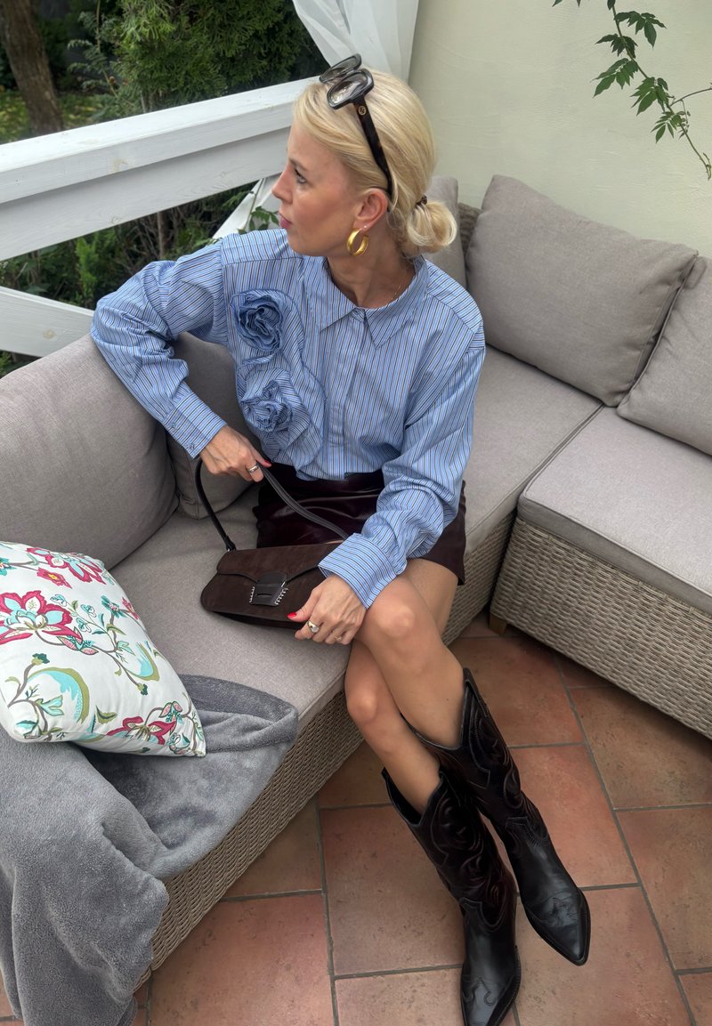Camisa de riscas azul de mangas compridas com detalhes florais, saia burgundy, botas de cowboy pretas e uma mala castanha sobre um sofá de tom claro.
