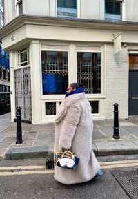 Manteau en fausse fourrure rose clair, ample avec un col montant. Sweat à capuche bleu dépassant. Porte un sac à main avec des accents blancs, bleus et beige.