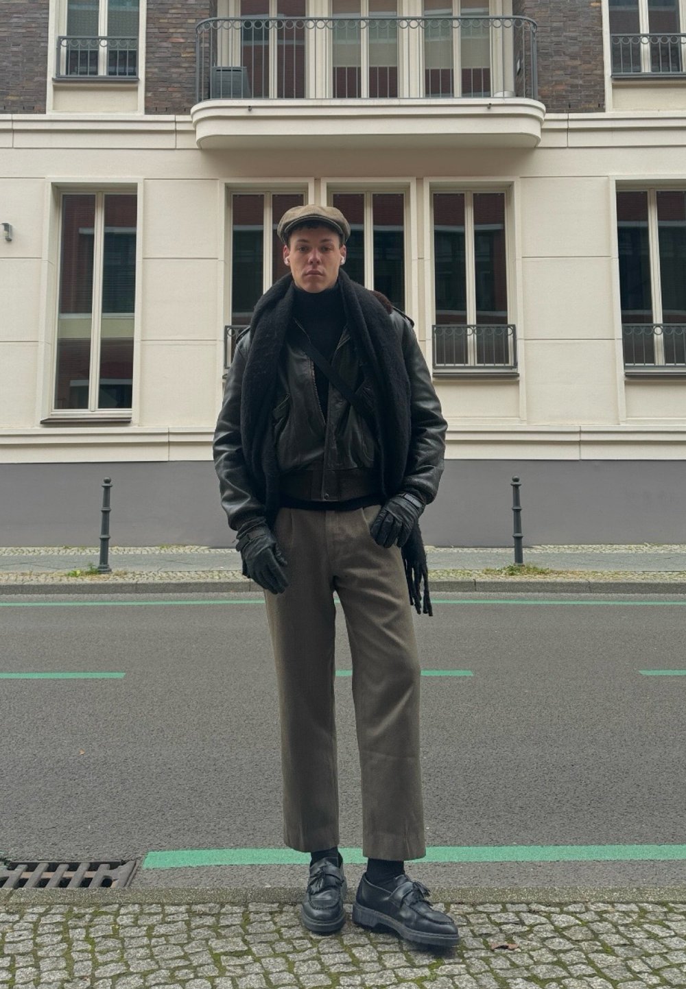 Veste en cuir noire, écharpe grise, gants noirs et casquette plate. Pantalons gris et chaussures noires. Cadre urbain avec une architecture moderne en arrière-plan.
