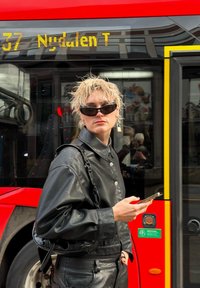 Conjunto de cuero negro con una chaqueta de botones y pantalones a juego. Los accesorios incluyen gafas de sol de ojo de gato y un pendiente metálico. Autobús rojo en el fondo.