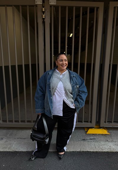 Persona sonriente de pie frente a una puerta metálica, usando chaqueta de mezclilla, cárdigan gris, pantalones blancos y negros, sosteniendo un bolso negro con una mano y con la otra en el bolsillo.