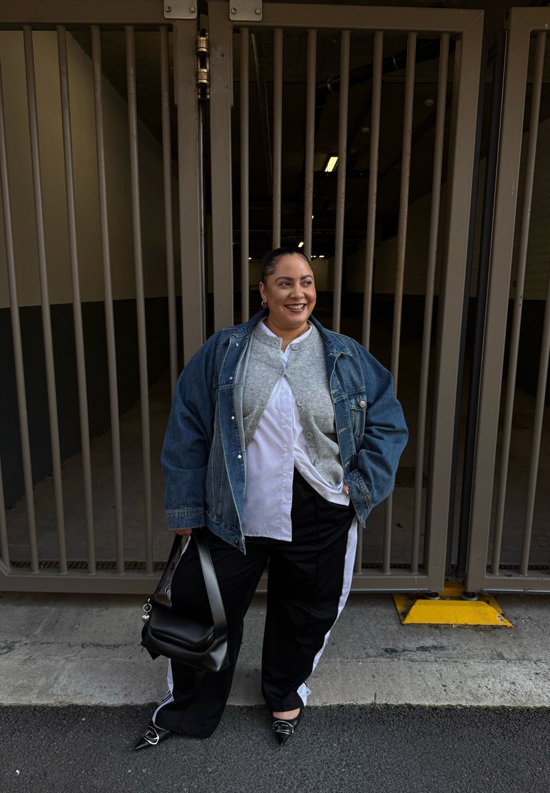 Personne souriante debout contre une grille métallique, portant une veste en jean, un cardigan gris, un pantalon noir et blanc, tenant un sac à main noir avec une main dans la poche.