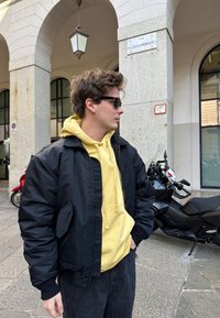 Črna bomberska jakna čez rumen kapuco, v kombinaciji z črnimi hlačami. Opozoriti je treba na teksturirano tkanino, sprednje žepe in sončna očala.