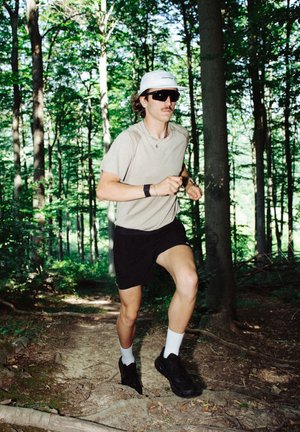 Corredor con una camisa de manga corta beige y pantalones cortos negros, que lleva gafas de sol y una gorra gris claro, corriendo por un sendero forestal entre árboles.