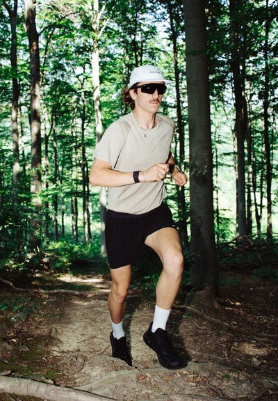 Corredor con una camisa de manga corta beige y pantalones cortos negros, que lleva gafas de sol y una gorra gris claro, corriendo por un sendero forestal entre árboles.