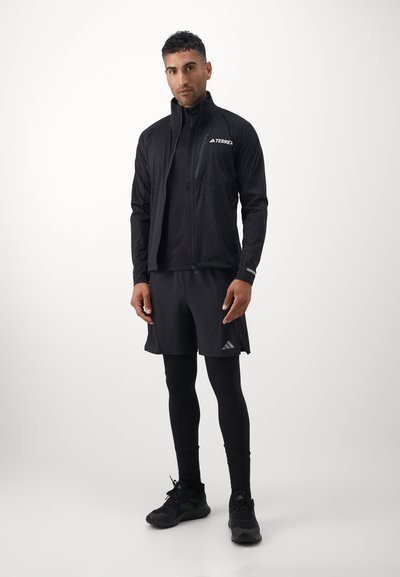 Chaqueta deportiva negra con cuello alto y cremallera, combinada con una camiseta negra ajustada y pantalones cortos. Presenta un logotipo sutil y una tela elástica.