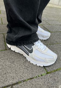 Τα παπούτσια τρεξίματος Nike διαθέτουν λευκό πλέγμα στο άνω μέρος με μαύρες ρίγες και λεπτομέρειες, παχιά λευκή σόλα και στρογγυλή σχεδίαση στα δάχτυλα. Φοριούνται μαύρα παντελόνια.