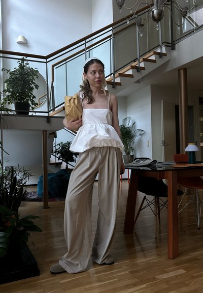 Top peplum blanco con tirantes finos, pantalones de pierna ancha beige con rayas y un bolso de mano dorado. Texturas suaves en un entorno interior moderno.