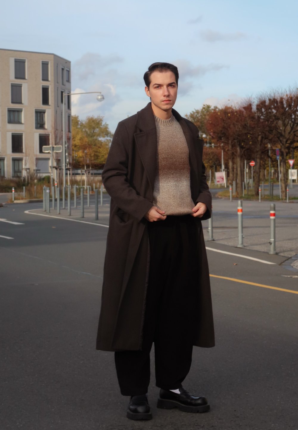 Lang, mørkebrun frakke over en lys beige sweater, sorte bukser og sorte sko. Udendørs bymiljø med grønne planter og bygninger i baggrunden.