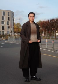 Lang, mørkebrun frakke over en lys beige sweater, sorte bukser og sorte sko. Udendørs bymiljø med grønne planter og bygninger i baggrunden.