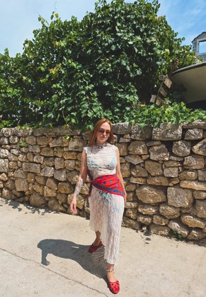 Vestido de encaje blanco, con una faja estampada en rojo y azul. Zapatos rojos. Fondo de pared de piedra, con follaje verde encima. Entorno soleado al aire libre.