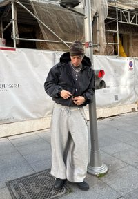 Veste noire courte avec capuche, pantalon de jogging gris à jambes larges, bottes noires et bonnet camouflage ; accessoirisé avec des chaînes en argent superposées.