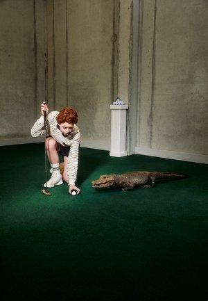 Una persona con un suéter a rayas se arrodilla en un césped verde, situada junto a un cocodrilo. Cerca, son visibles una pelota de golf y un palo.