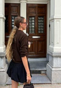 Suéter de malha castanho, saia plissada preta, óculos de sol, segurando uma bebida. Cabelo longo com um elástico, em pé perto de portas de madeira com painéis de vidro.