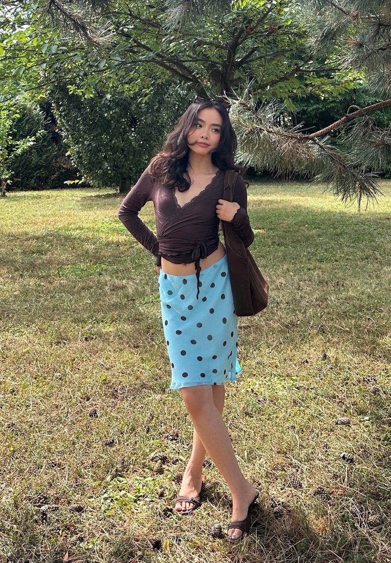Woman standing on grass under tree branches, wearing brown long-sleeve top, blue polka dot skirt, sandals, and carrying a brown bag.