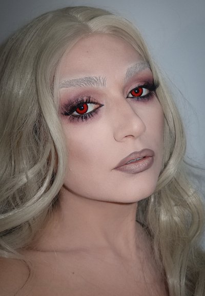 Cabello largo y rubio claro con textura ondulada. Piel pálida con acabado mate. El maquillaje incluye sombra de ojos oscura, lentes de contacto rojos en la esclerótica y un color de labios apagado.