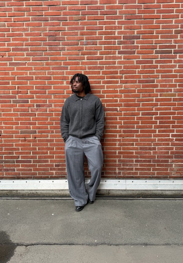 Mężczyzna z dredami, ubrany w ciemną kurtkę na zamek i szerokie szare spodnie, stoi z rękami w kieszeniach przy czerwonej ceglanej ścianie na betonowym podłożu.