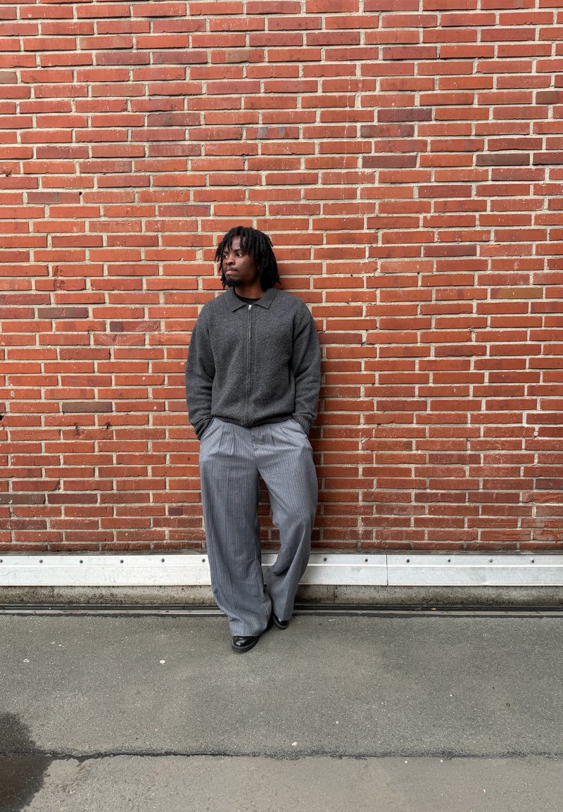 Homme aux dreadlocks portant une veste à fermeture éclair foncée et un pantalon large gris, debout les mains dans les poches contre un mur en briques rouges sur un sol en béton.