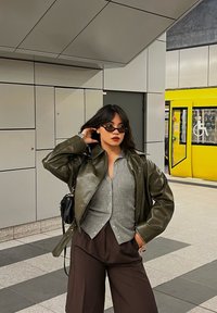 Žena u sunčanim naočalama, zelenoj kožnoj jakni, sivoj majici i smešnim hlačama stoji na železničkoj stanici s žutim vlakom iza nje.