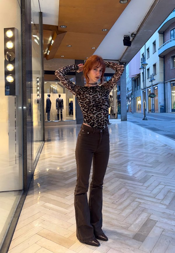 Jonge vrouw in een top met luipaardprint en donkere broek poseert met haar handen achter haar hoofd in een moderne winkelgalerij.