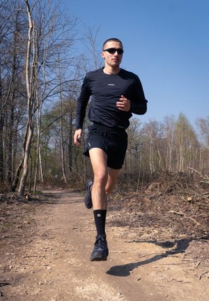 Hombre vestido con ropa deportiva negra y gafas de sol corriendo por un sendero de tierra a través de un bosque sin hojas bajo un cielo azul despejado.