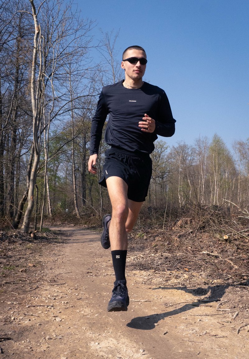 Man wearing black sportswear and sunglasses running on a dirt trail through a leafless forest under clear blue sky.