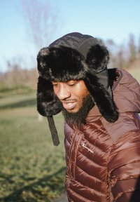 Chapeau de trappeur doublé de fourrure noire avec rabats d'oreilles, porté avec une veste isolante marron. Textures douces, couleurs contrastantes visibles en plein air.