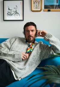 Homme barbu portant une écharpe colorée, allongé sur un canapé bleu, vêtu d'un pull gris et de bagues, avec une œuvre encadrée sur un mur blanc en arrière-plan.