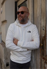 Felpa bianca con tasca frontale e logo sul petto, indossata con jeans scuri. Occhiali da sole rotondi, marroni. Porta di legno texturizzata sullo sfondo.