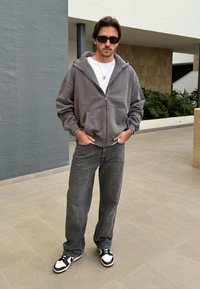 Sweatshirt cinza com fecho, sobre uma t-shirt branca, combinado com jeans cinzas de corte largo e ténis preto e branco. Os acessórios incluem óculos de sol e pulseiras.