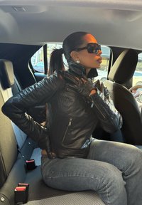 Schwarze Lederjacke mit strukturiertem Finish, mit Reißverschluss versehenen Taschen und schwarzen Handschuhen. Sitzt in einem Auto mit grauen Sitzen und sichtbaren Gurtschlössern.