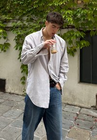 Übergroßes, hellgrau gestreiftes Hemd mit Knöpfen; blaue Jeans mit weitem Bein; hält ein Glas mit Strohhalm, gefüllt mit Eiskaffee. Grüne Blätter im Hintergrund.