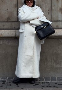 Personne portant un long manteau blanc, une écharpe blanche, un chapeau blanc, de grandes lunettes de soleil noires, des bottes noires, tenant un sac à main noir contre un mur de pierre.