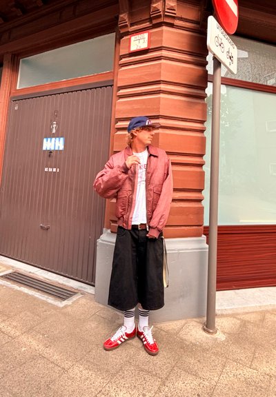 Persona con una chaqueta bomber color burdeos, camiseta blanca, culottes negros, zapatillas rojas y calcetines rayados se encuentra junto a un edificio con molduras decorativas.