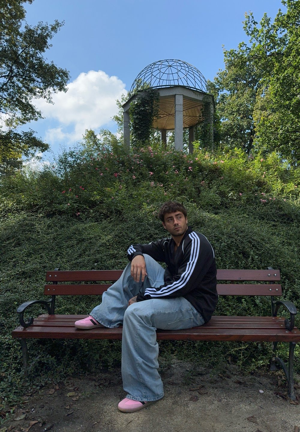 Άτομο με μαύρο μπουφάν με άσπρες ρίγες, φαρδιά ανοιχτό μπλε τζιν και ροζ παπούτσια, καθισμένο σε ένα καφέ παγκάκι στο πάρκο ανάμεσα σε πράσινα φυτά και λουλούδια.