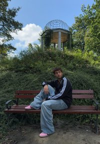 Άτομο με μαύρο μπουφάν με άσπρες ρίγες, φαρδιά ανοιχτό μπλε τζιν και ροζ παπούτσια, καθισμένο σε ένα καφέ παγκάκι στο πάρκο ανάμεσα σε πράσινα φυτά και λουλούδια.