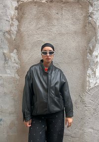 Schwarze Leder-Übergröße-Jacke mit hohem Kragen, kombiniert mit schwarzen Hosen mit silbernen Ösen, vor einer strukturierten grauen Wand.