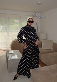 Vestido de bolinhas pretas e brancas com mangas longas e decote com laço. Acessorado com óculos de sol oversized e uma pequena mala preta.