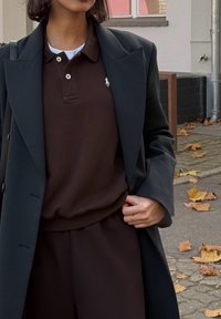 Barna galléros póló fekete kabát alatt, kis hímzett logóval, laza szabással és jól látható textúrával. A barna nadrág teszi teljessé az összeállítást.