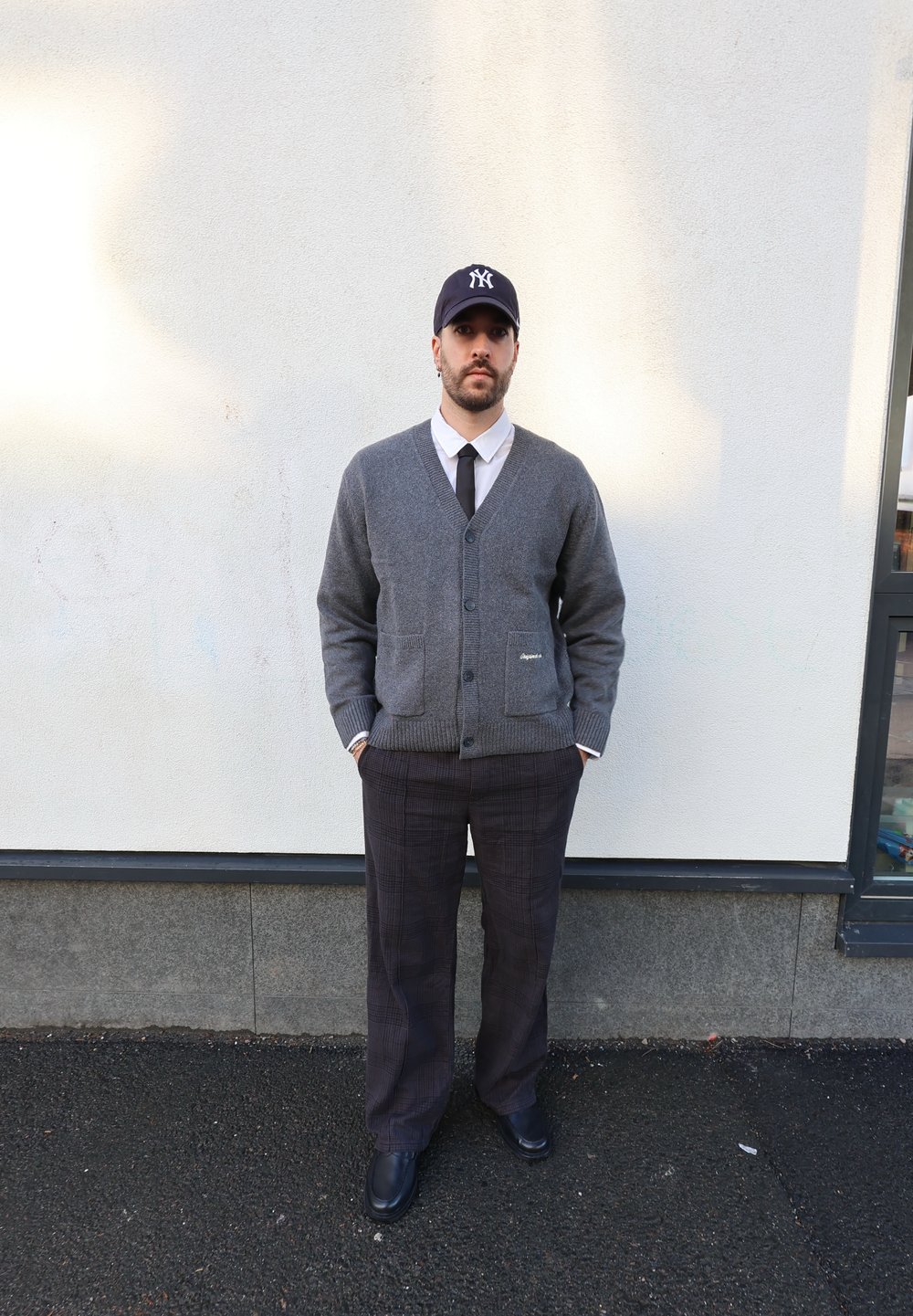 Cardigan en tricot gris sur une chemise blanche avec une cravate noire, assorti à un pantalon à carreaux, des chaussures noires et une casquette de baseball bleu marine.