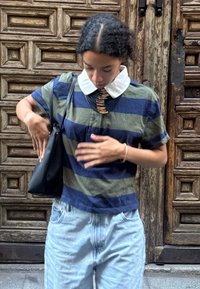 Persona vistiendo una camiseta a rayas en tonos navy y olivo con un cuello blanco ajusta una bolsa negra de hombro frente a una puerta de madera detallada.