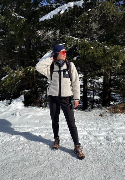 Chaqueta de forro polar con parte frontal negra, textura esponjosa gris claro, pantalones negros y botas de senderismo marrones. Gafas de sol de colores brillantes y gorro de lana. Fondo nevado.