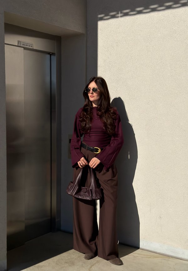 Mujer con gafas de sol, una camiseta de manga larga morada oscura y pantalones anchos marrones, sosteniendo un bolso marrón oscuro, de pie cerca de un ascensor a la luz del sol.