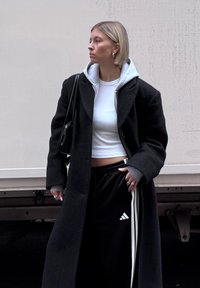 Donna con capelli biondi corti che indossa un cappotto nero sopra un felpa grigia e un top bianco corto, pantaloni da jogging neri Adidas e porta una borsa a tracolla nera.