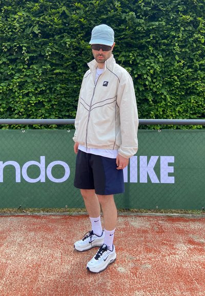 Hombre con chaqueta ligera, pantalones cortos negros, gorra y gafas de sol de pie en una pista delante de una valla verde con logotipos de marcas deportivas.
