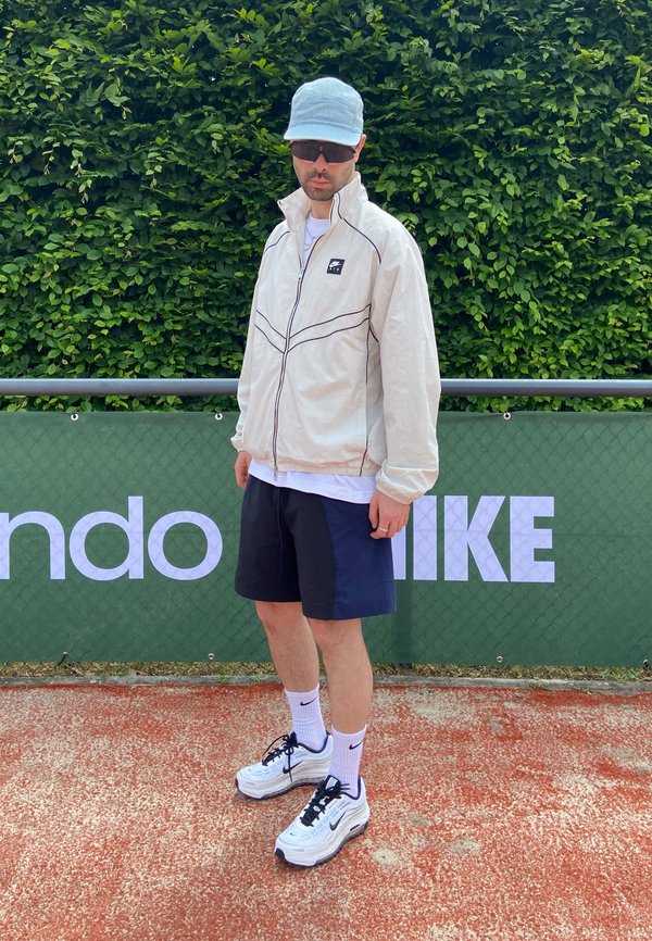 Hombre con chaqueta ligera, pantalones cortos negros, gorra y gafas de sol de pie en una pista delante de una valla verde con logotipos de marcas deportivas.