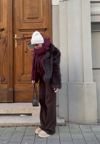 Femme en tenue d'hiver avec un bonnet blanc, une écharpe bordeaux, un manteau en fourrure sombre et un pantalon marron, debout sur un pavage en pierre près d'une porte en bois.